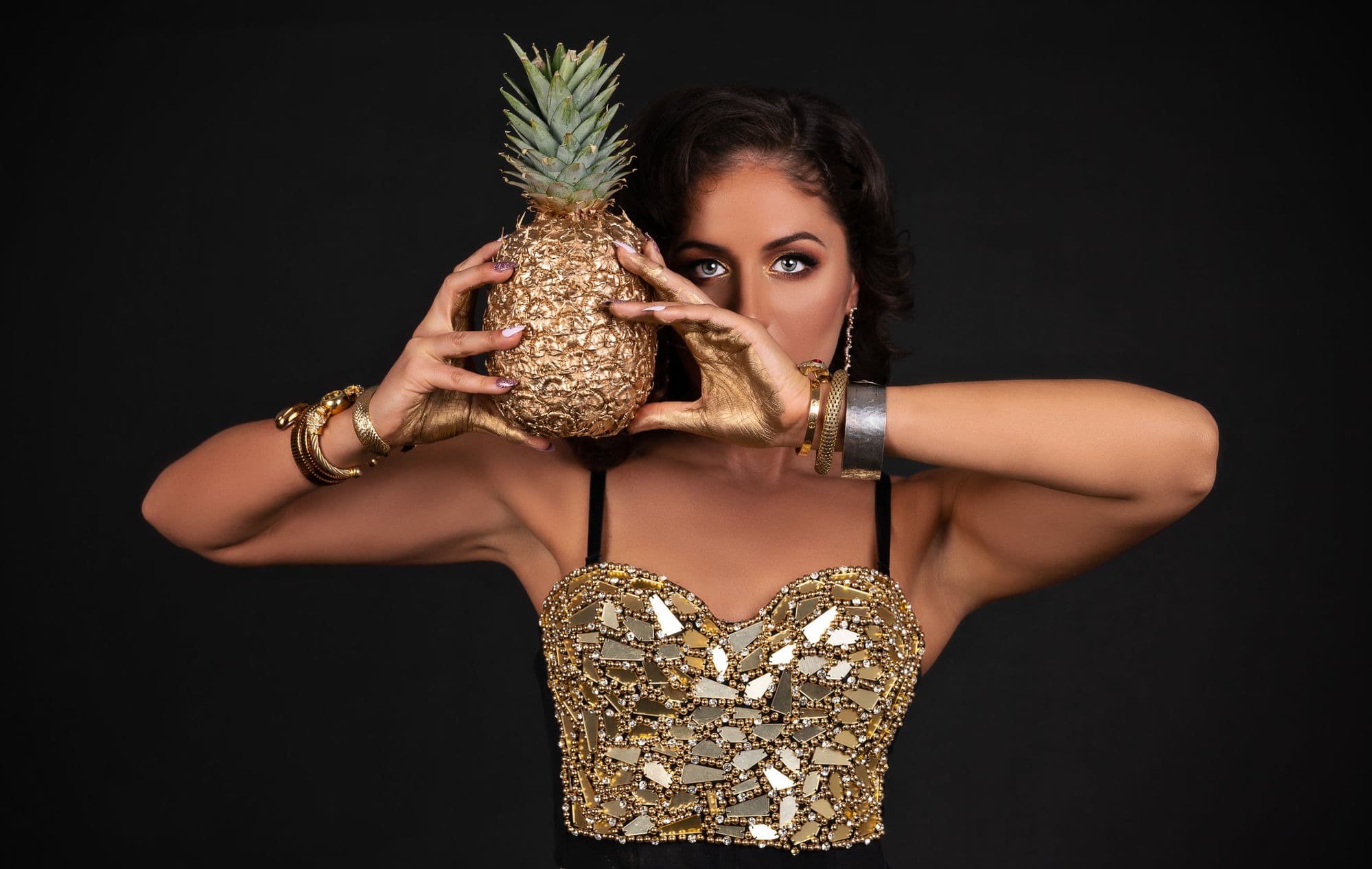 An editorial beauty shot of a woman in a gold bustier holding a gilded pineapple against black